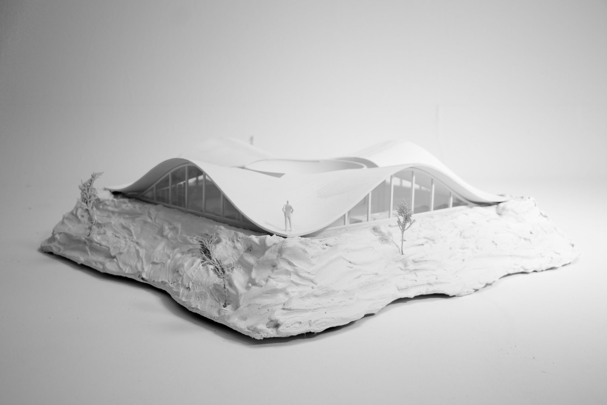 White model of a house under one wavy roof that rises and dips, set on a sculpted hill, with a small figure standing on the roof edge.