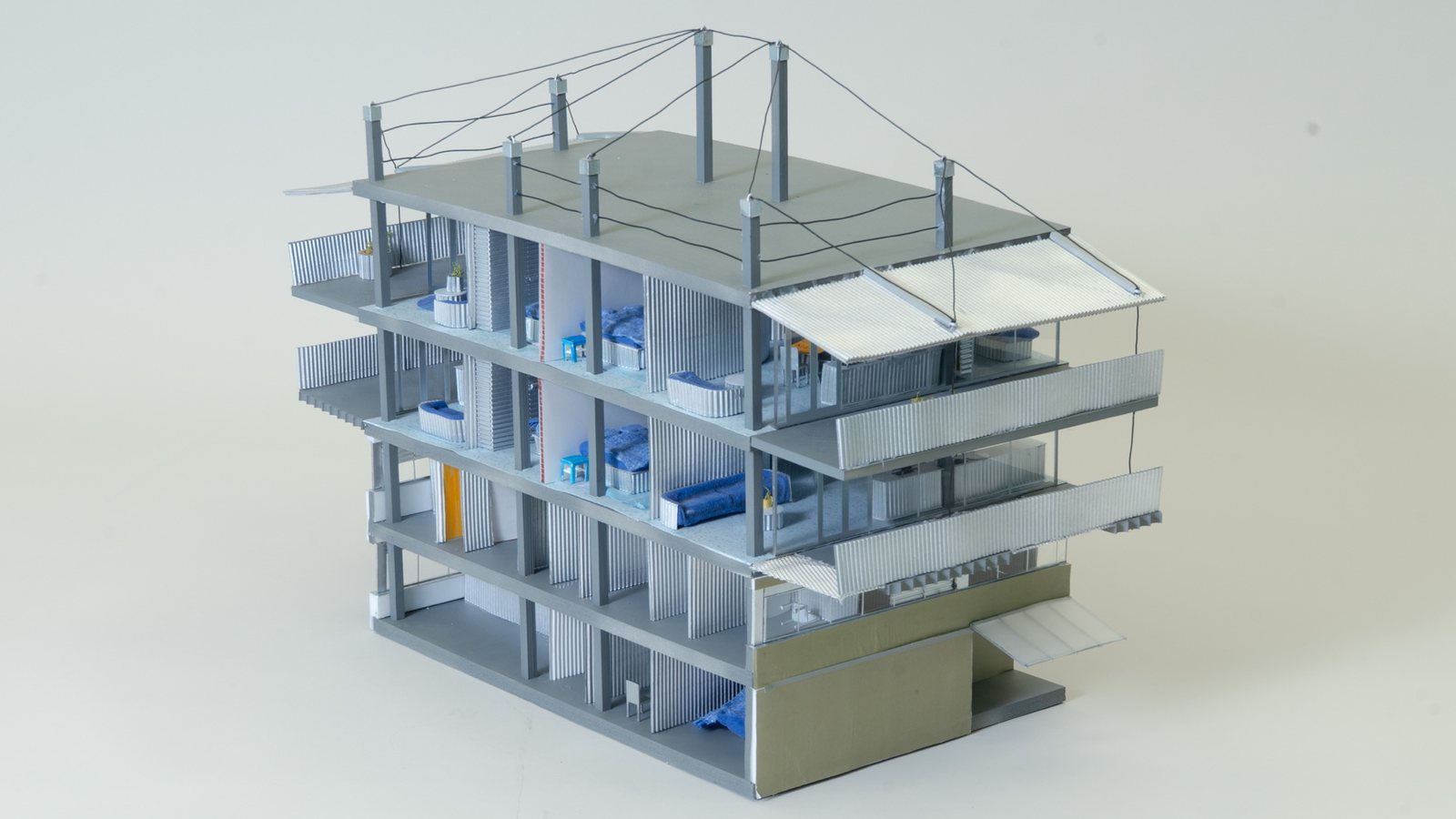 Grey card model of a four-storey building cut open to show rooms with blue furnishings, balconies with corrugated screens and posts with cables on the roof.